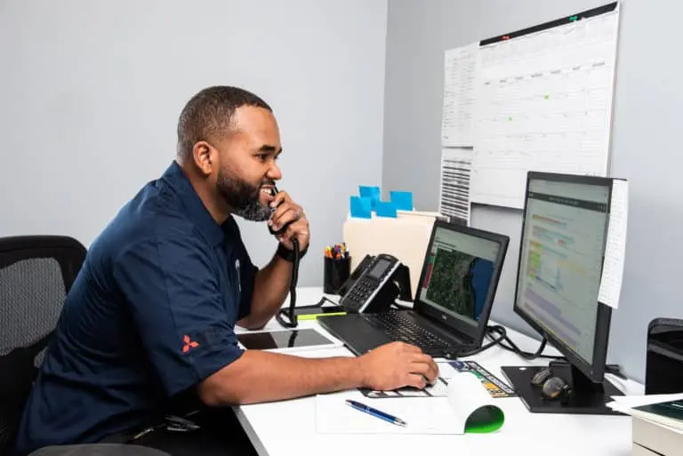 A man with a beard in a blue polo shirt sits at a white desk, talking on a landline phone while using a computer mouse. Two monitors, a laptop and a vertical external display, are visible on the desk, along with office supplies and documents.