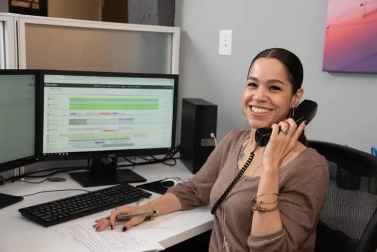 A smiling woman sits at an office desk, talking on a landline phone while looking at dual computer monitors and holding a pen over a document.
