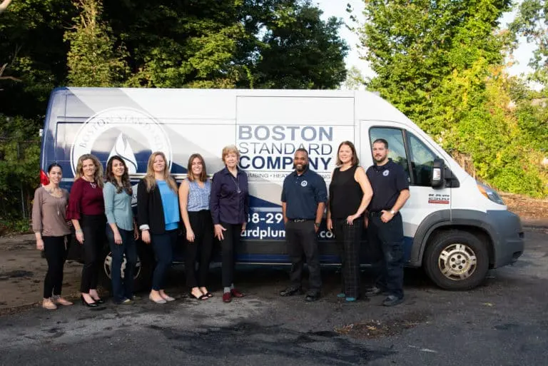 A group of nine people, including seven women and two men, stand in front of a white and blue service van with 'Boston Standard Company' written on its side. They are on an asphalt surface with green trees in the background.