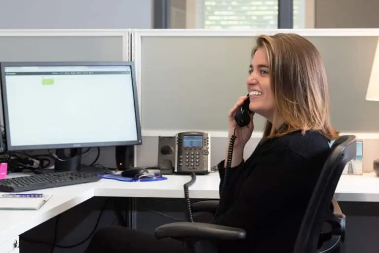 A smiling woman sits at a desk in an office cubicle, talking on a landline phone. A computer monitor and keyboard are on the desk.