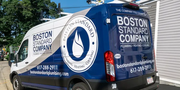 A white and blue service van for Boston Standard Company, featuring their logo with a flame icon and text for plumbing, heating, and cooling services. Contact information is visible on the side and back.