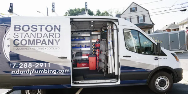 A white and blue service van with its side door open, revealing an organized interior with shelves holding various plumbing and HVAC tools and equipment, including a red toolbox and ladders. The van's side is branded with "BOSTON STANDARD COMPANY PLUMBING HEATING COOLING".