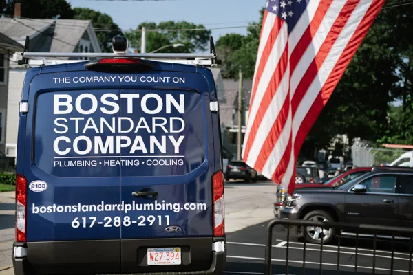 Rear view of a blue Boston Standard Company service van with its branding, website, and phone number, parked next to a large American flag.