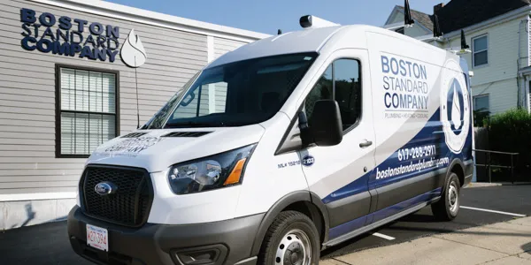 A white Boston Standard Company service van with blue and grey branding is parked in front of a building displaying the company's name.