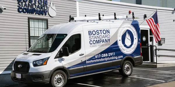 A white service van with blue and gray branding for Boston Standard Company, a plumbing, heating, and cooling business, is parked in front of a gray-sided building. An American flag hangs near the building's entrance.
