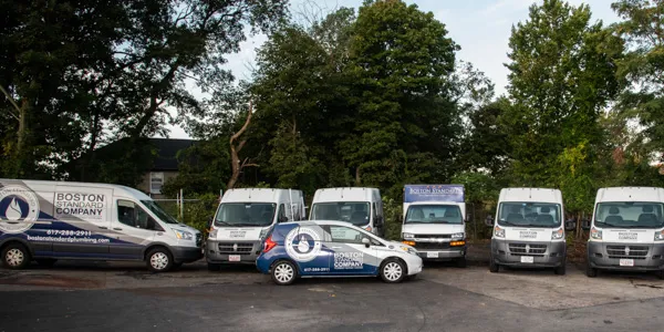 A fleet of white service vans and one car, branded with 'Boston Standard Company' and a logo, parked in a lot surrounded by trees.