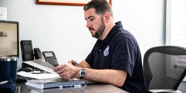 A man with a beard sits at an office desk, looking at a document. A computer monitor, phone, and tumbler are on the desk.