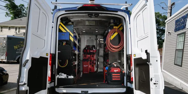 Rear view of an open white utility van, revealing a meticulously organized interior with shelving units, various tools, coiled hoses, and storage bins for equipment. The van is parked on a street with buildings in the background.