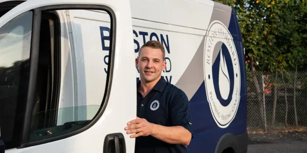 A smiling male service technician in a dark blue polo shirt stands by the open door of a white service van branded "BOSTON SEWER & DRAIN".