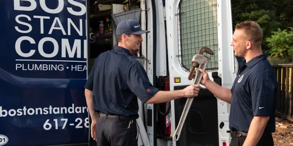 Two service technicians in dark blue uniforms stand by an open service van, one handing a large pipe wrench to the other.