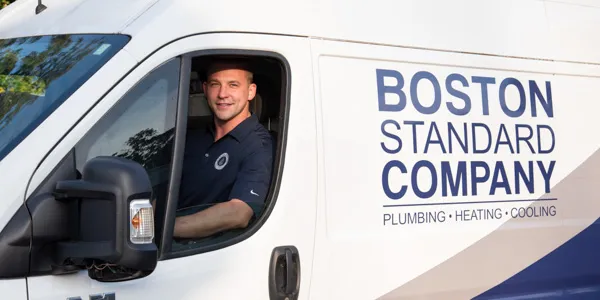 A smiling man in a dark polo shirt sits in the driver's seat of a white service van, which displays 'BOSTON STANDARD COMPANY PLUMBING • HEATING • COOLING' in blue text on its side.