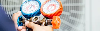 A person holds an HVAC manifold gauge set with blue and red dials in front of an air conditioning unit, exploring finance options for upgrading their cooling system.