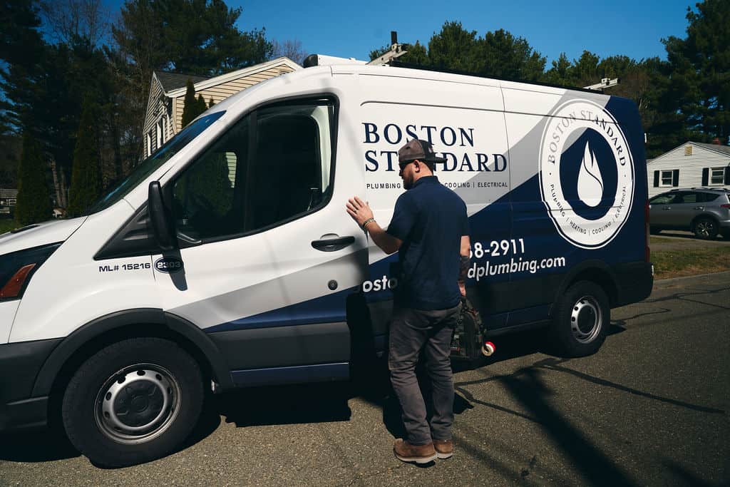 A person wearing a cap stands next to a white Boston Standard plumbing van parked on a suburban street.