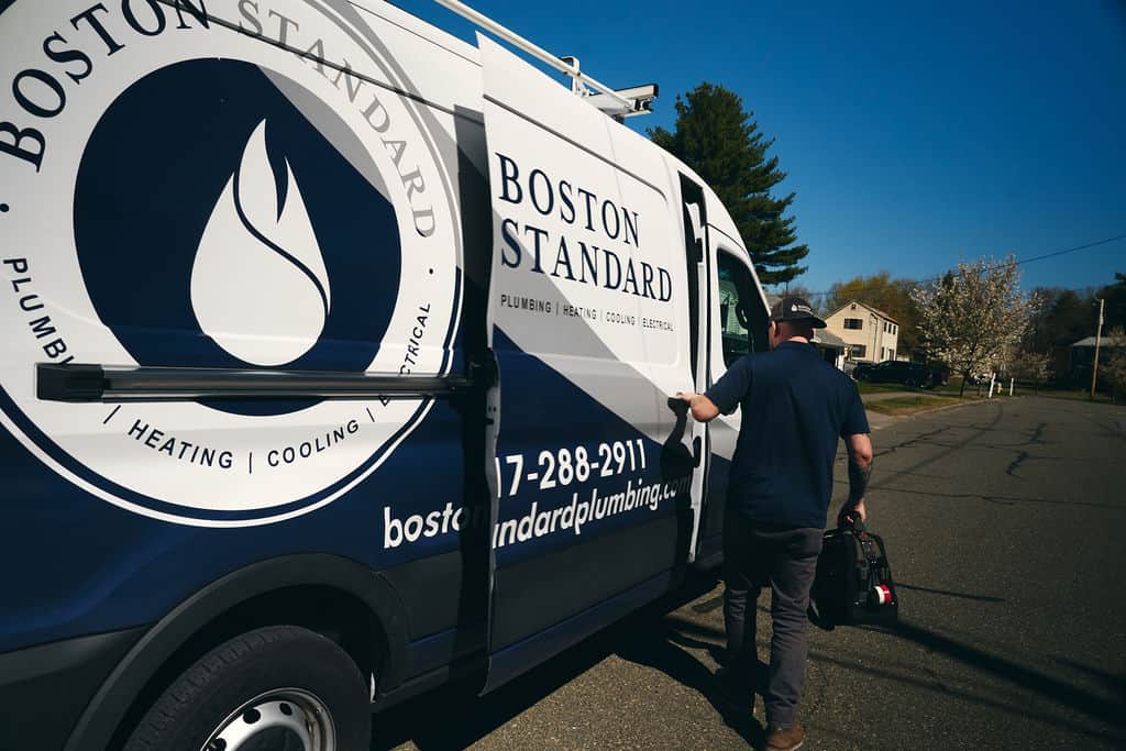 A person carrying a tool bag walks toward a Boston Standard plumbing service van parked on a suburban street.