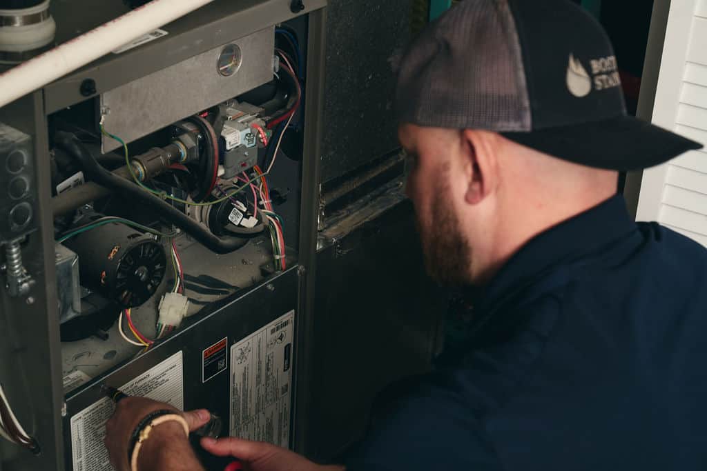 A man wearing a cap repairs an HVAC unit, inspecting wiring and components inside the open panel, as part of professional heating services Boston residents rely on.