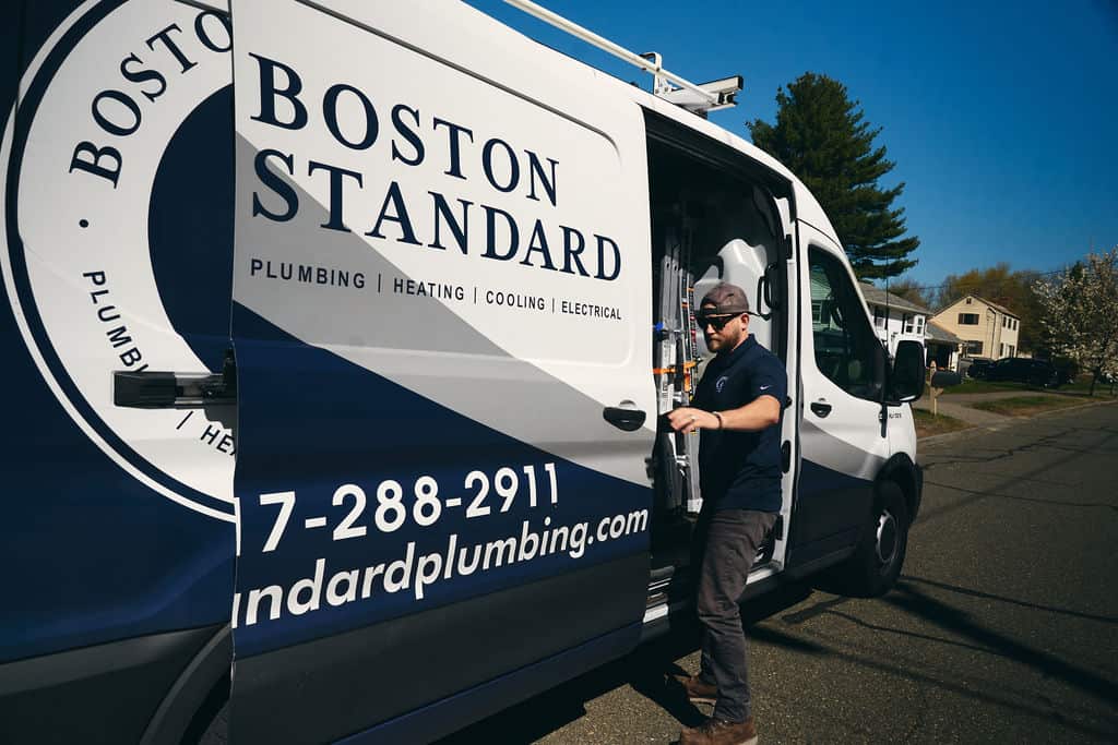 A plumber stands beside an open Boston Standard company van, preparing for a job on a sunny residential street.