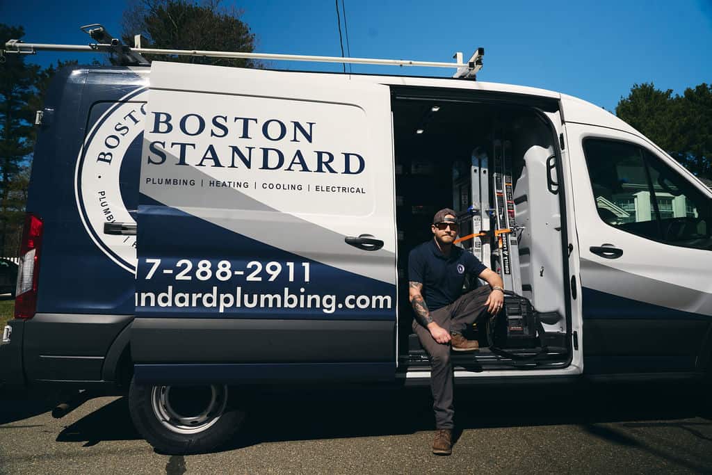 A plumber sits on the edge of an open Boston Standard service van, with tools and equipment visible inside, on a sunny day.
