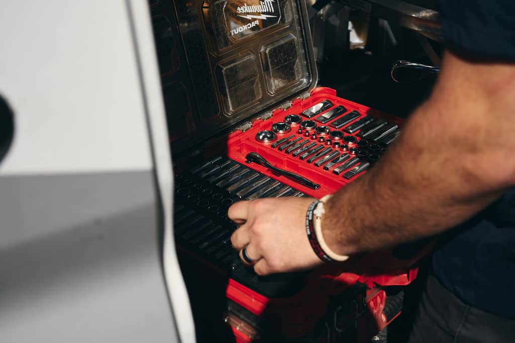 A person selects a tool from a red socket set case containing various sockets, a ratchet, and other hand tools—essential equipment for generator safety and surge protection during storms.